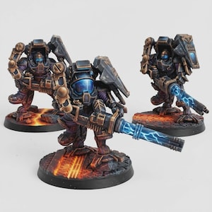 May include: Three miniature figures of armored soldiers with futuristic weapons. The figures are painted in dark blue, bronze, and black, with glowing blue energy effects emanating from their weapons. Each figure stands on a circular base.