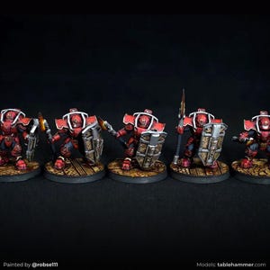 May include: Five miniature figurines of armoured warriors. Each figure is painted in red and black with gold accents, holding weapons and shields. The figures stand on circular bases with a textured, brown surface, set against a dark background.