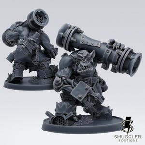 Op de afbeelding: Twee grijze miniatuurfiguren van Orcs, één knielend en de ander staand, die elk grote, cilindrische wapens dragen. De figuren zijn gedetailleerd met pantser en mechanische componenten, geplaatst op ronde bases. Het Smuggler Boutique-logo is zichtbaar.