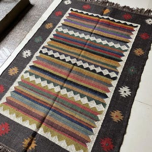 May include: Rectangular area rug with a dark gray border and a central design of colorful horizontal stripes. The stripes include shades of red, yellow, blue, and green. The rug has a fringed edge and decorative floral and geometric patterns.