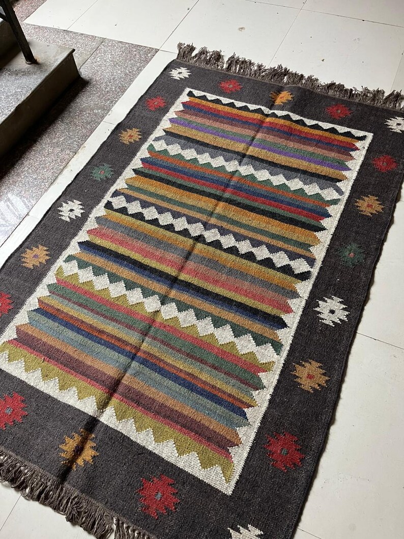 May include: Rectangular area rug with a dark gray border and a central design of colorful horizontal stripes. The stripes include shades of red, yellow, blue, and green. The rug has a fringed edge and decorative floral and geometric patterns.