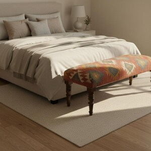 May include: A bedroom scene featuring a bed with pillows and a patterned bench at the foot. The bench has a wooden frame and a colorful, geometric fabric. The room has a neutral color palette with a rug and hardwood flooring.