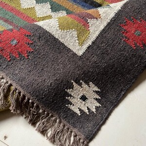 May include: A handwoven rug with a geometric pattern and fringed edges. The rug features a dark gray border with red floral accents and a central design of white, green, and yellow geometric shapes. The rug's main body has stripes of various colors.