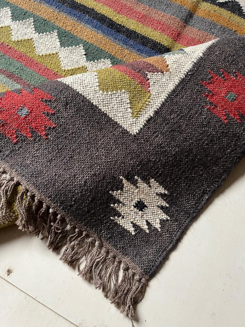 May include: A handwoven rug with a geometric pattern and fringed edges. The rug features a dark gray border with red floral accents and a central design of white, green, and yellow geometric shapes. The rug's main body has stripes of various colors.
