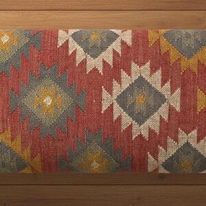 May include: Rectangular bench cushion with a geometric pattern in red, yellow, gray, and white. The cushion has a woven texture and is placed on a wooden surface. The design features diamond and zigzag shapes.