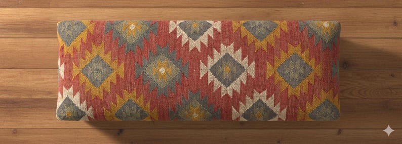 May include: Rectangular bench cushion with a geometric pattern in red, yellow, gray, and white. The cushion has a woven texture and is placed on a wooden surface. The design features diamond and zigzag shapes.