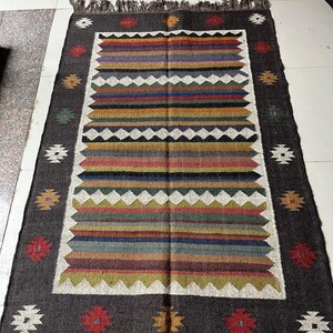 May include: Rectangular rug with a dark gray border and a central design of horizontal stripes in various colors, including red, blue, green, and yellow. The rug has a fringed edge and decorative floral patterns in the border.