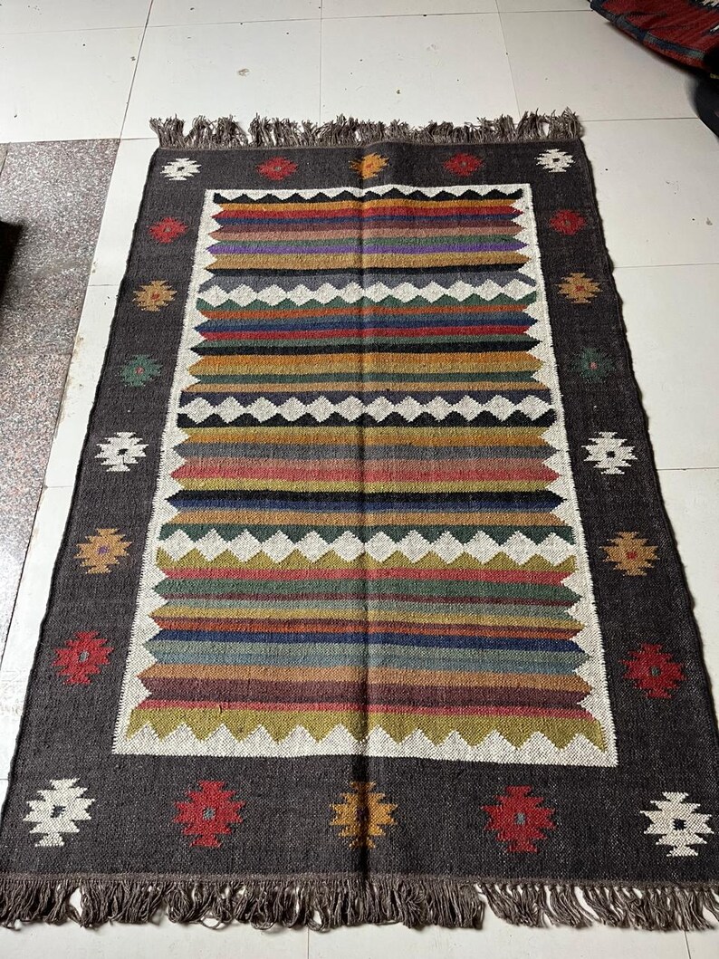 May include: Rectangular rug with a dark gray border and a central design of horizontal stripes in various colors, including red, blue, green, and yellow. The rug has a fringed edge and decorative floral patterns in the border.