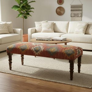 May include: A rectangular bench with a patterned fabric top in shades of red, orange, yellow, and gray. The bench has dark brown, turned legs and is placed on a neutral-colored rug in a living room setting.