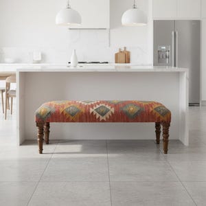 May include: A decorative bench with a patterned fabric seat in shades of red, yellow, and gray. The bench has four turned wooden legs. The bench is placed in front of a white kitchen island.