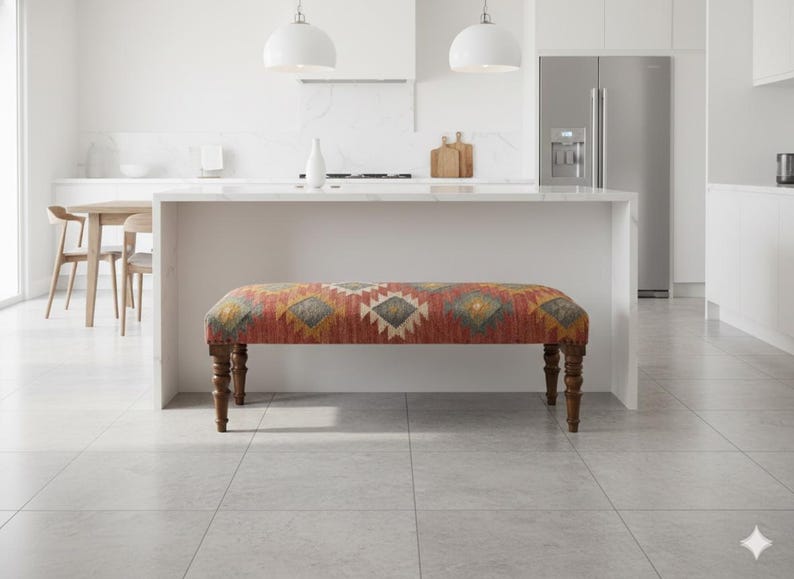 May include: A decorative bench with a patterned fabric seat in shades of red, yellow, and gray. The bench has four turned wooden legs. The bench is placed in front of a white kitchen island.