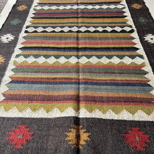 May include: Rectangular woven rug with a dark gray border and a central design of horizontal stripes in various colors, including red, yellow, and blue. The rug features a fringed edge and floral patterns.