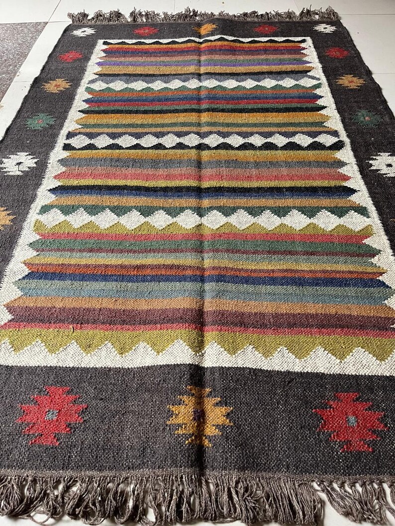May include: Rectangular woven rug with a dark gray border and a central design of horizontal stripes in various colors, including red, yellow, and blue. The rug features a fringed edge and floral patterns.
