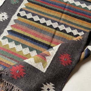May include: A hand-woven rug with a dark gray border and a central design of colorful horizontal stripes. The stripes include shades of red, orange, yellow, green, blue, and purple. The rug has a fringed edge and floral patterns in the corners.