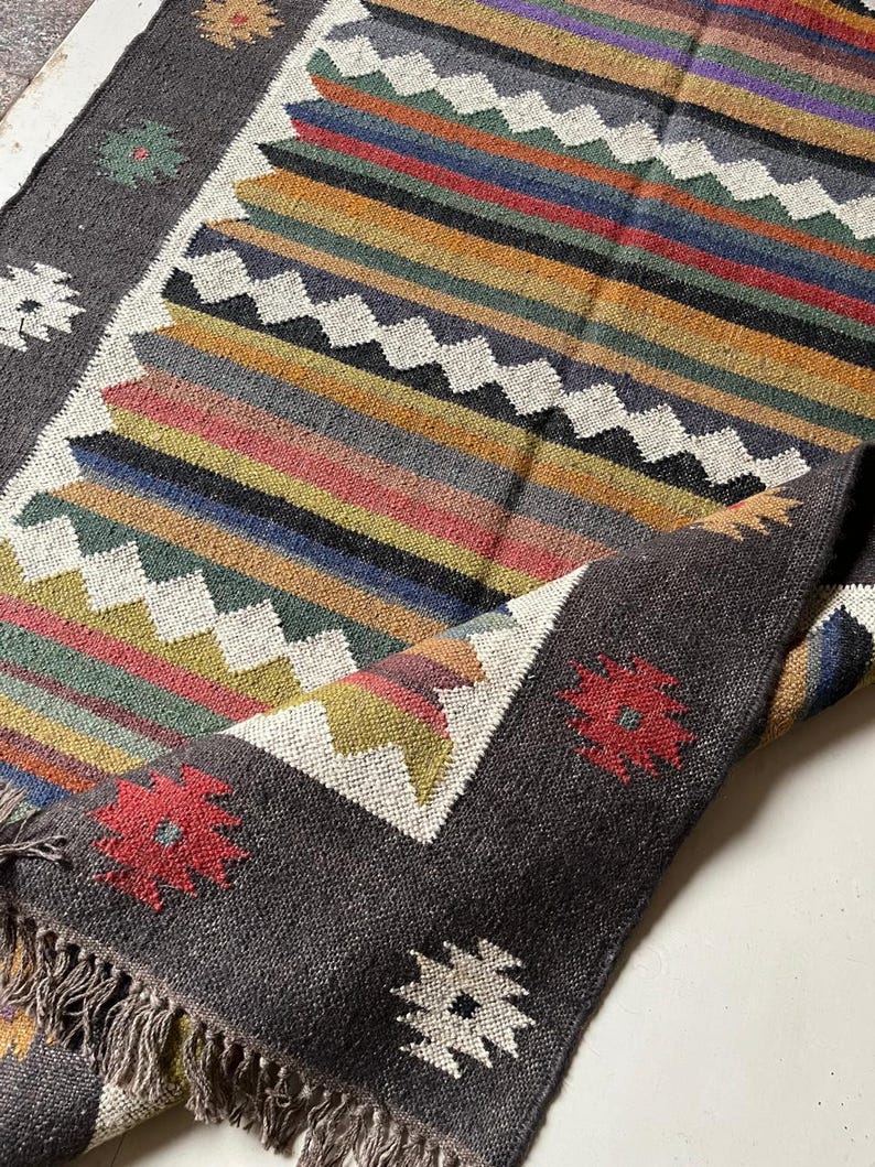 May include: A hand-woven rug with a dark gray border and a central design of colorful horizontal stripes. The stripes include shades of red, orange, yellow, green, blue, and purple. The rug has a fringed edge and floral patterns in the corners.