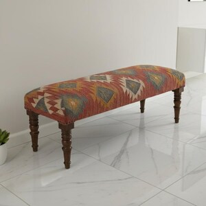 May include: Upholstered bench with a patterned design in red, yellow, and cream colors. The bench has dark brown wooden legs. The bench is a decorative furniture piece.