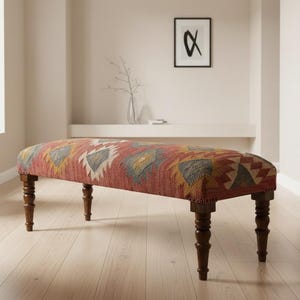May include: An upholstered bench with a red, orange, yellow, and gray geometric pattern. The bench has dark brown, turned legs. The bench is in a room with light-colored walls and hardwood floors.
