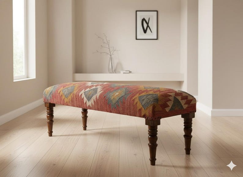 May include: An upholstered bench with a red, orange, yellow, and gray geometric pattern. The bench has dark brown, turned legs. The bench is in a room with light-colored walls and hardwood floors.