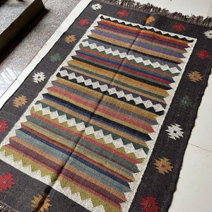 May include: A rectangular, hand-woven rug with a dark gray border and a central design of colorful horizontal stripes. The stripes include shades of red, blue, green, and yellow. The rug has a fringed edge and decorative floral and geometric patterns.