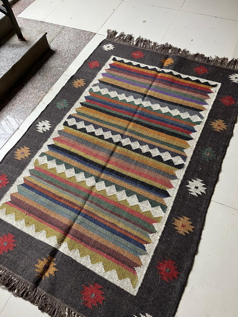 May include: A rectangular, hand-woven rug with a dark gray border and a central design of colorful horizontal stripes. The stripes include shades of red, blue, green, and yellow. The rug has a fringed edge and decorative floral and geometric patterns.