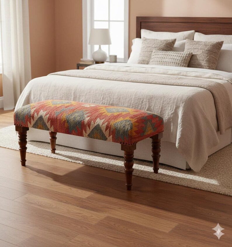 May include: A decorative bench with a patterned fabric seat in shades of red, orange, yellow, and gray. The bench has four dark wooden legs and is placed at the foot of a bed with neutral-colored bedding. The room has a warm, inviting atmosphere.