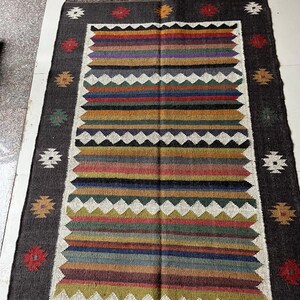 May include: Rectangular rug with a dark gray border and a central design of horizontal stripes in various colors, including red, orange, green, and blue. The rug has a white zigzag pattern and decorative floral accents.