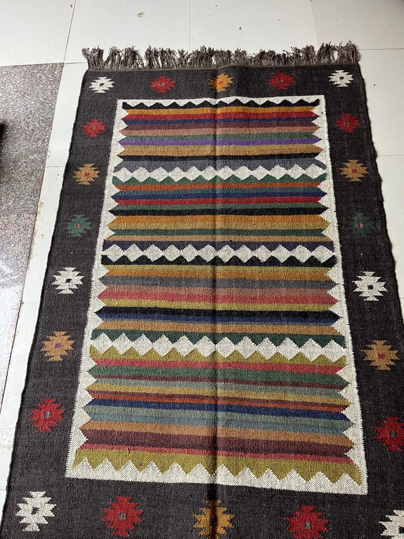 May include: Rectangular rug with a dark gray border and a central design of horizontal stripes in various colors, including red, orange, green, and blue. The rug has a white zigzag pattern and decorative floral accents.