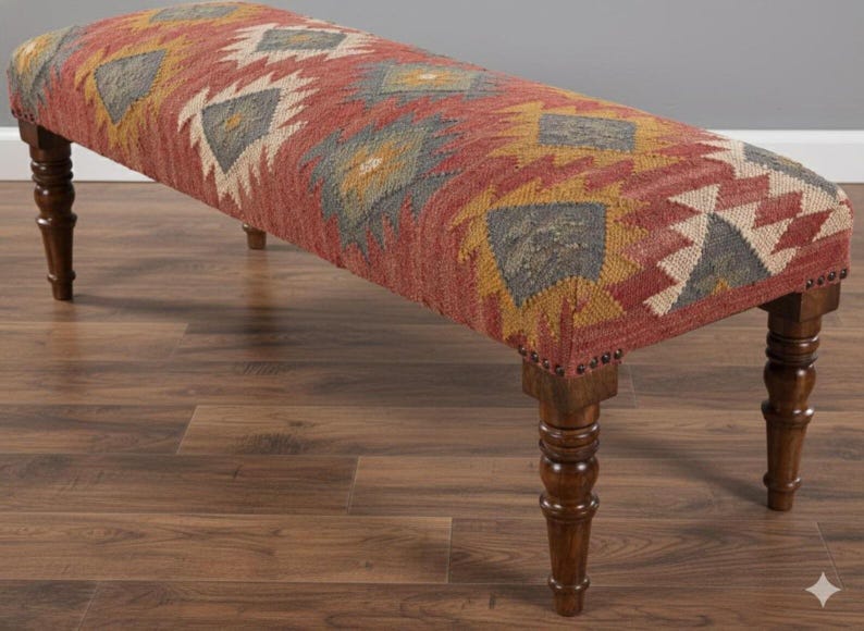 May include: A long, upholstered bench with a patterned top in shades of red, yellow, gray, and white. The bench has turned wooden legs and is suitable for a living room or entryway. The bench measures approximately 48 inches long.