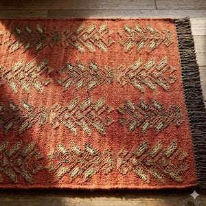 May include: Terracotta-colored rug with a repeating geometric pattern in shades of beige and brown. The rug has a textured surface and a fringed edge. The design features stylized leaf-like shapes, creating a warm and inviting aesthetic.