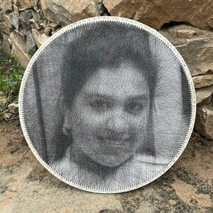 Può includere: Opera d'arte murale rotonda con un ritratto creato con filo nero. L'opera è montata su una cornice circolare con bordi bianchi. Lo sfondo è un muro di pietra naturale.