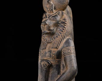 May include: A dark gray statue of a deity with a lion's head and a sun disc headdress. The statue has intricate carvings on the chest, arms, and headdress. The figure is standing and has a serene expression. The background is black.
