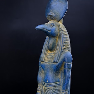 May include: A blue statue of an Egyptian deity with a bird head and a sun disc. The statue has intricate details, including a patterned headdress and clothing. The figure is set against a dark background.