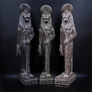 Peut inclure: Trois statues en pierre d'une divinité à tête de lion, coiffée d'une coiffe et d'un collier orné. Les statues sont dans des tons de gris et de marron, sur des socles rectangulaires avec des hiéroglyphes. Le fond est noir.