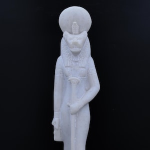 May include: A white sculpture of a deity with a lion's head, wearing a headdress with a sun disc. The figure holds a staff and a small pouch. The statue has detailed carvings and stands against a black background.