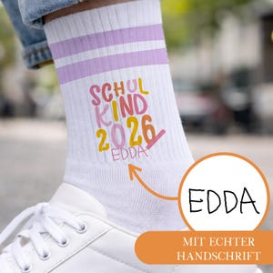 Op de afbeelding: Een witte sok met twee lichtpaarse strepen aan de bovenkant. De sok is voorzien van de kleurrijke tekst SCHUL KIND 2026 en de naam EDDA in een handgeschreven stijl. Een oranje pijl wijst naar de naam met een cirkelvormig inzetstuk EDDA.