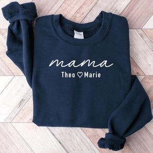May include: A navy blue sweatshirt featuring the word "mama" in white script. Underneath, the text reads "Theo ♡ Marie" in white. The garment is folded on a light brown wooden surface.