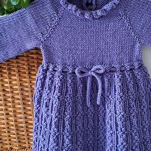 May include: A lavender-colored knitted dress with long sleeves. The dress features a ruffled collar, a decorative bow at the waist, and a textured skirt. The dress is displayed on a black hanger.