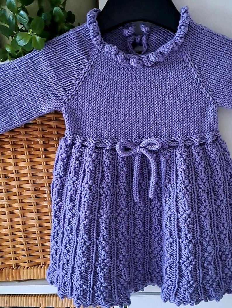 May include: A lavender-colored knitted dress with long sleeves. The dress features a ruffled collar, a decorative bow at the waist, and a textured skirt. The dress is displayed on a black hanger.