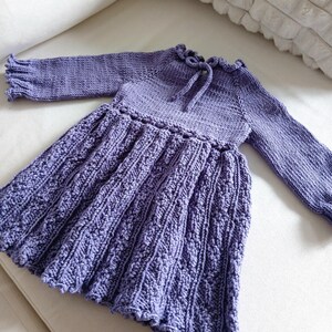 May include: A lavender-colored knitted dress with long sleeves. The dress features a textured skirt, a ruffled neckline with a tie, and ruffled sleeve cuffs. The dress is made of soft yarn and is suitable for a baby or toddler.