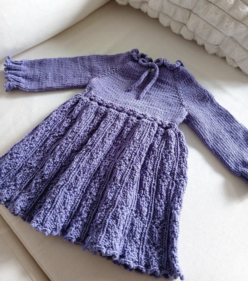 May include: A lavender-colored knitted dress with long sleeves. The dress features a textured skirt, a ruffled neckline with a tie, and ruffled sleeve cuffs. The dress is made of soft yarn and is suitable for a baby or toddler.