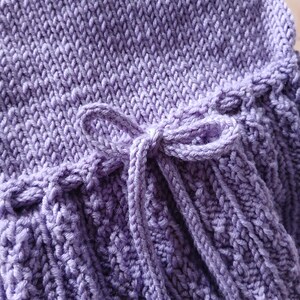May include: Close-up of a lavender knitted item with a textured pattern. The item features a decorative bow made from the same yarn. The knitted fabric has a ribbed design and a soft, inviting appearance.