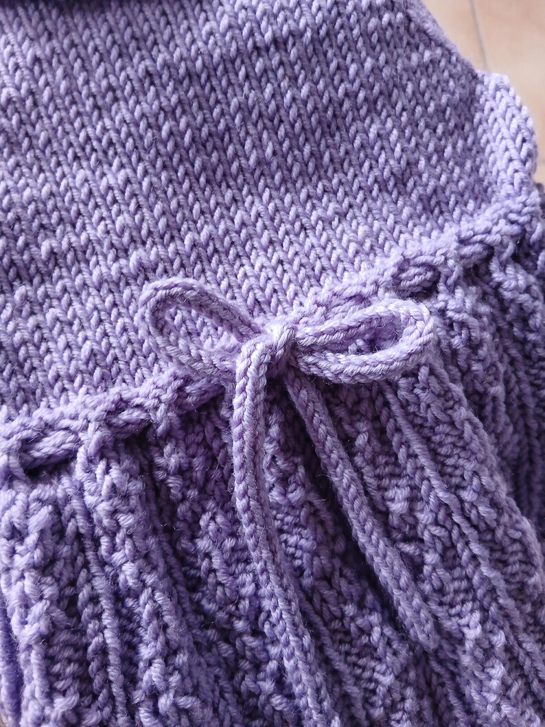 May include: Close-up of a lavender knitted item with a textured pattern. The item features a decorative bow made from the same yarn. The knitted fabric has a ribbed design and a soft, inviting appearance.