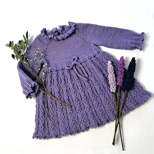 May include: A lilac-colored knitted dress with long sleeves and a ruffled neckline. The dress features a textured skirt and a tie at the waist. Decorative sprigs of lavender and greenery are arranged near the dress, creating a soft, feminine aesthetic.