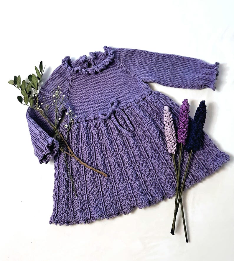 May include: A lilac-colored knitted dress with long sleeves and a ruffled neckline. The dress features a textured skirt and a tie at the waist. Decorative sprigs of lavender and greenery are arranged near the dress, creating a soft, feminine aesthetic.
