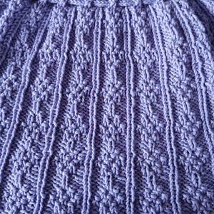 May include: Close-up of a lavender knitted garment. The textured fabric features vertical ribbed sections and intricate diamond patterns. The garment has a scalloped edge and a detailed, braided waistband.