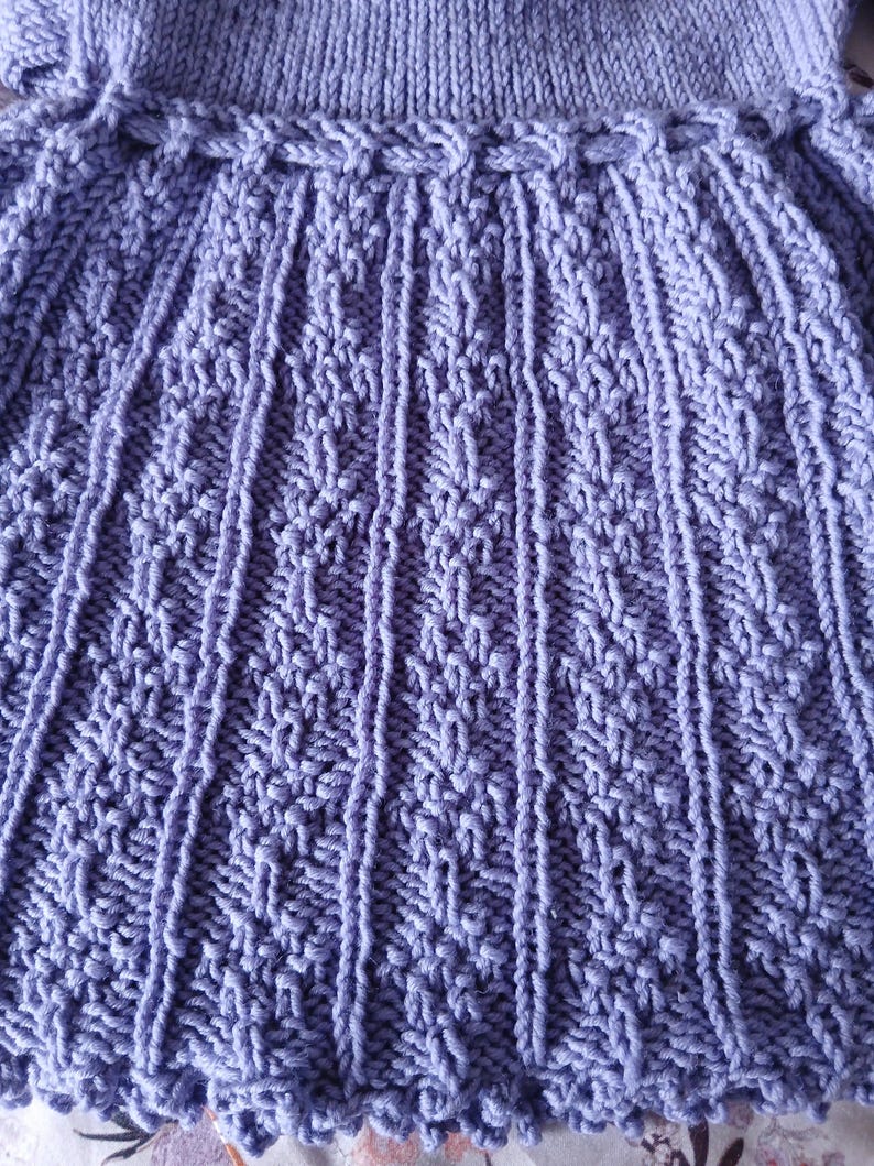 May include: Close-up of a lavender knitted garment. The textured fabric features vertical ribbed sections and intricate diamond patterns. The garment has a scalloped edge and a detailed, braided waistband.