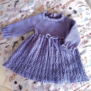May include: A lavender-colored, hand-knitted dress with long sleeves and a ruffled neckline. The skirt features a textured pattern and a tie at the waist. The dress is designed for a child.