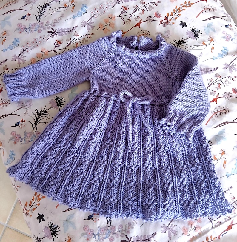 May include: A lavender-colored, hand-knitted dress with long sleeves and a ruffled neckline. The skirt features a textured pattern and a tie at the waist. The dress is designed for a child.