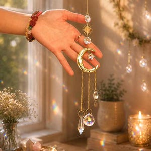 May include: A gold-toned metal suncatcher featuring a crescent moon charm, a small star, and multiple clear faceted crystal prisms suspended by delicate chains. The decorative hanging ornament includes a sunburst emblem and round crystal beads.