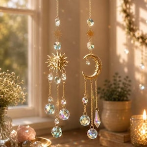 May include: Two hanging suncatchers featuring gold-toned metal sun and crescent moon charms. Each piece is suspended by a delicate gold chain and adorned with multiple clear, multifaceted crystal beads that refract light into colourful prisms.